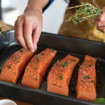 A cook is sprinkling herbs on four pieces of raw salmon.